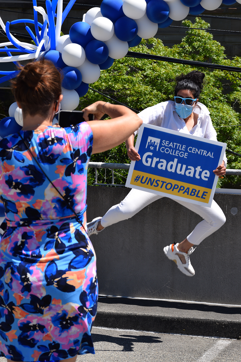 a female graduate jumps as her teacher takes her picture under a white and blue balloon arch